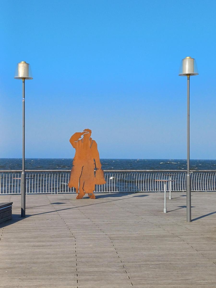 Holzplattform mit Metallgeländer, Skulptur und Blick auf Meer unter blauem Himmel.