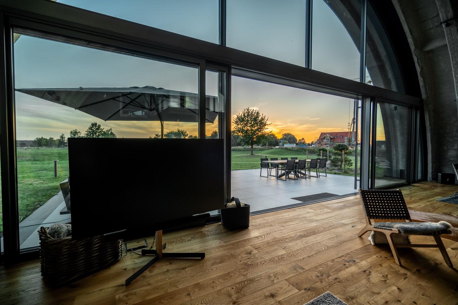 Große Glasfront mit Blick auf Terrasse und Sonnenuntergang. Holzboden und Sitzmöbel im Raum.