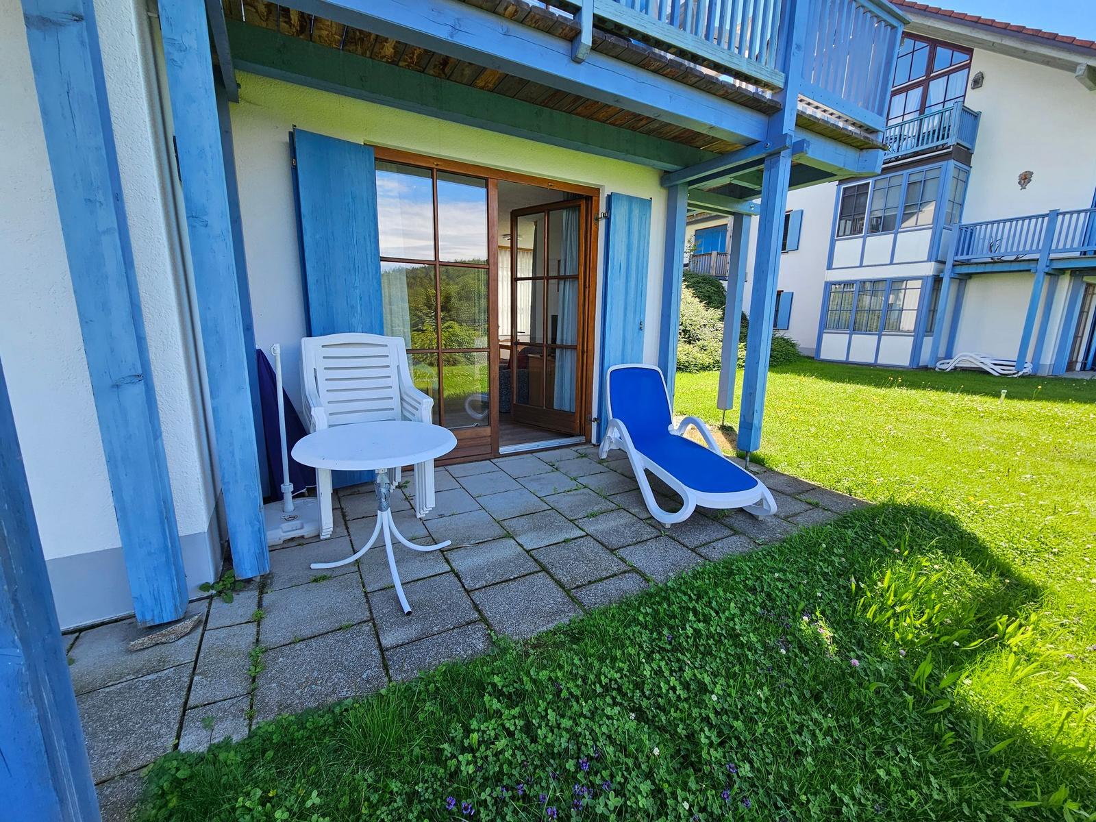 Terrasse mit Tisch, Stuhl und Liegestuhl. Hintergrund: grüner Rasen und Gebäude.