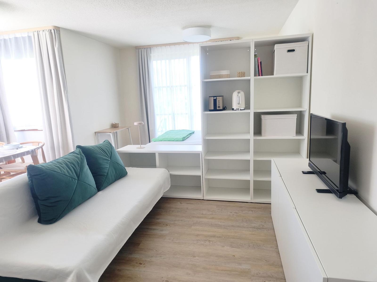 Wohnraum mit Sofa, Regal, Fernseher und Fenster mit Vorhängen.