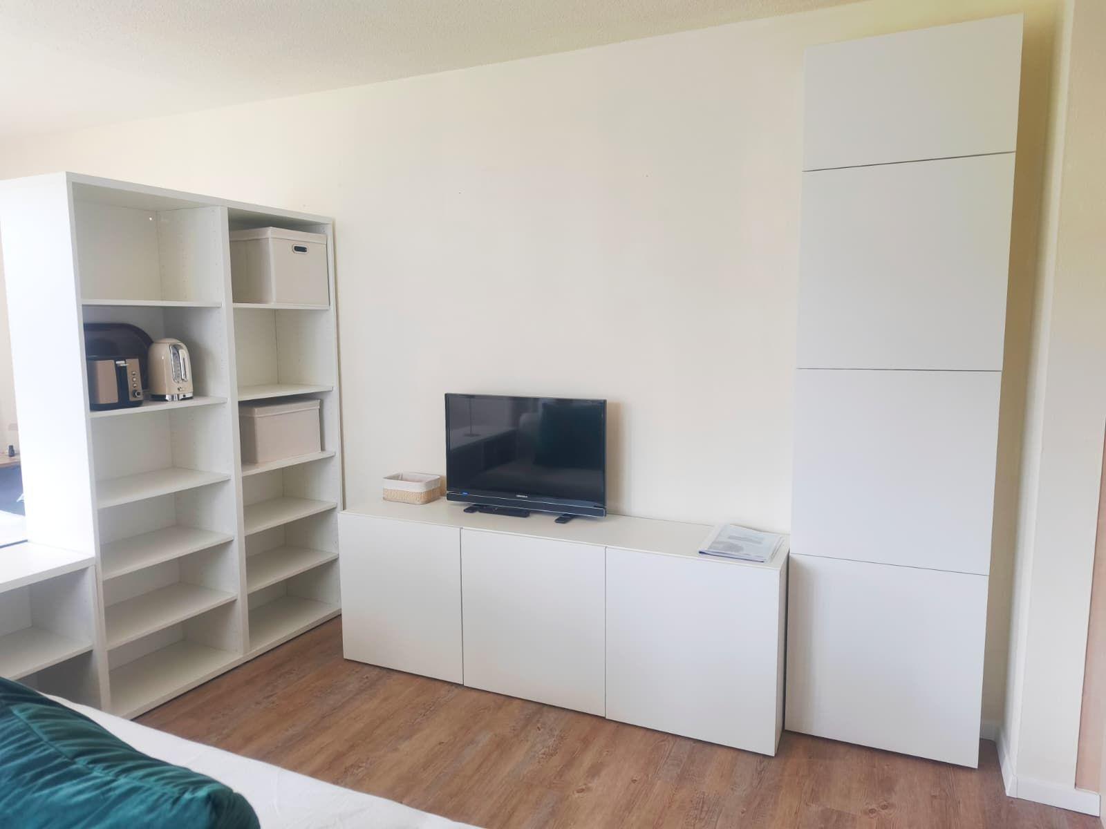 Zimmer mit weißen Schränken, Fernseher und Küchenutensilien auf Regalen.