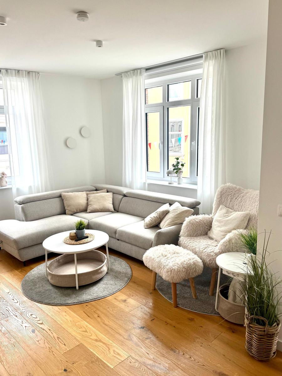 Wohnzimmer mit Ecksofa, Rundtisch und Fellstuhl vor großen Fenstern.