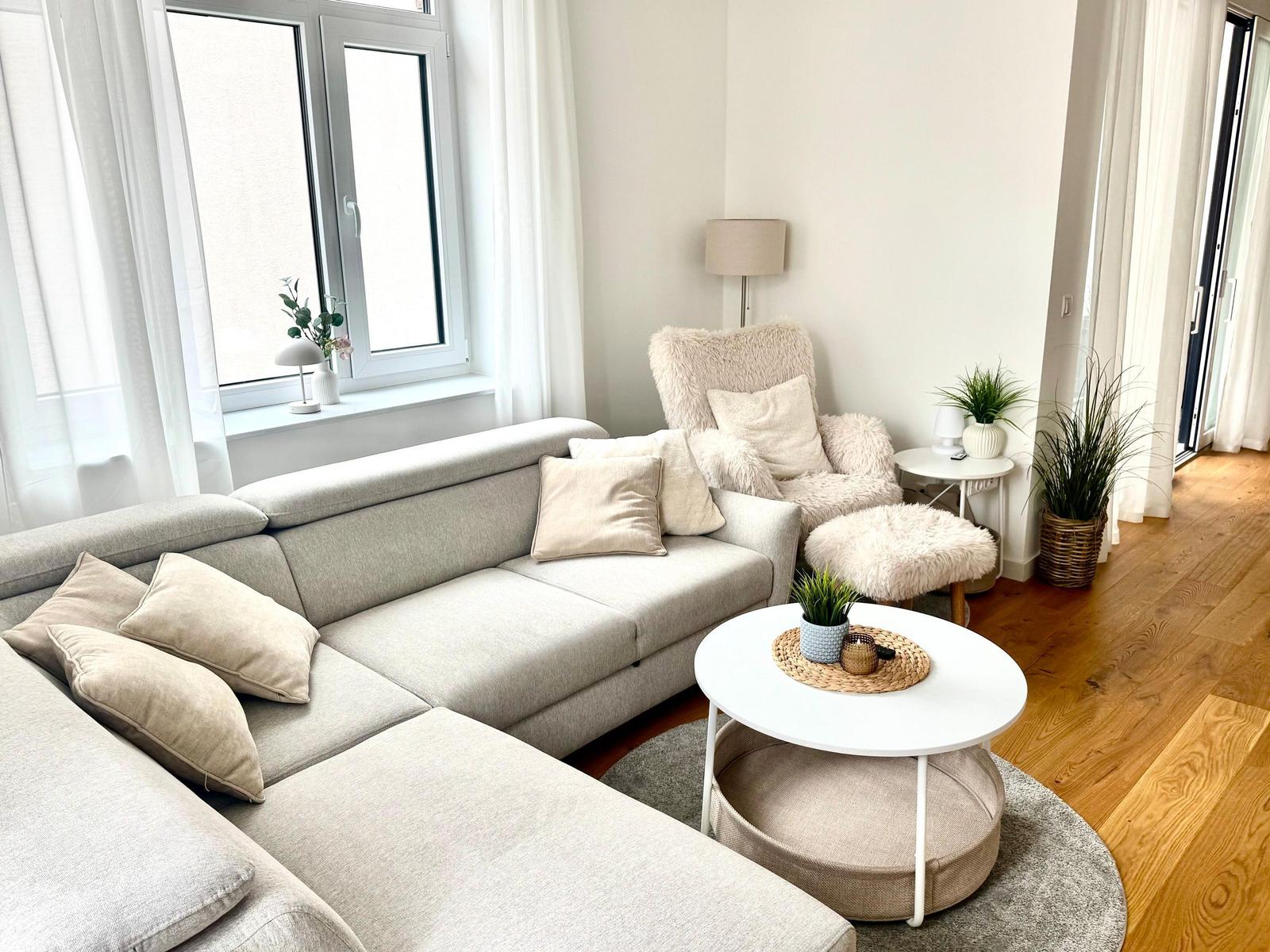 Ein Wohnzimmer mit grauem Sofa, runden Tisch und Sessel. Holzboden und Fenster mit weißen Vorhängen.