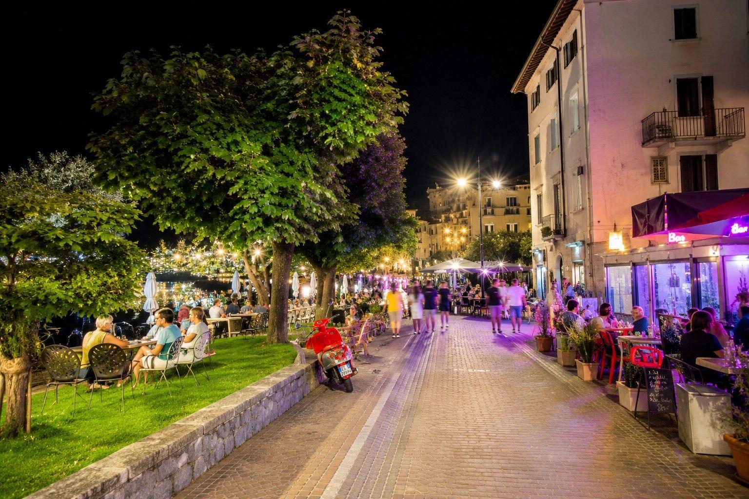 Nächtliche Straße mit Restaurants, Bäumen und einem roten Roller.