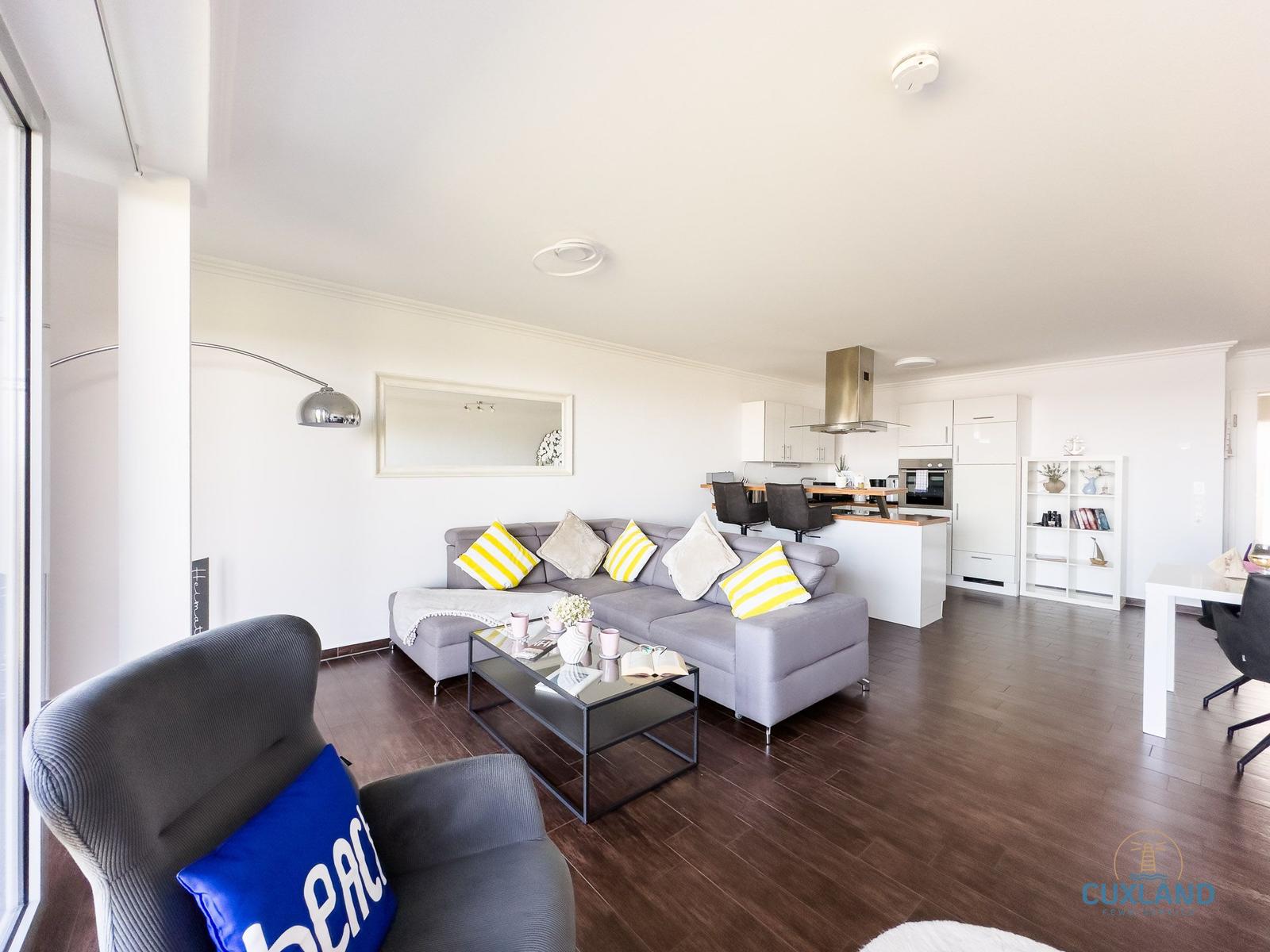 Wohn- und Küchenbereich mit grauem Sofa, gelben Kissen und dunklem Holzboden.