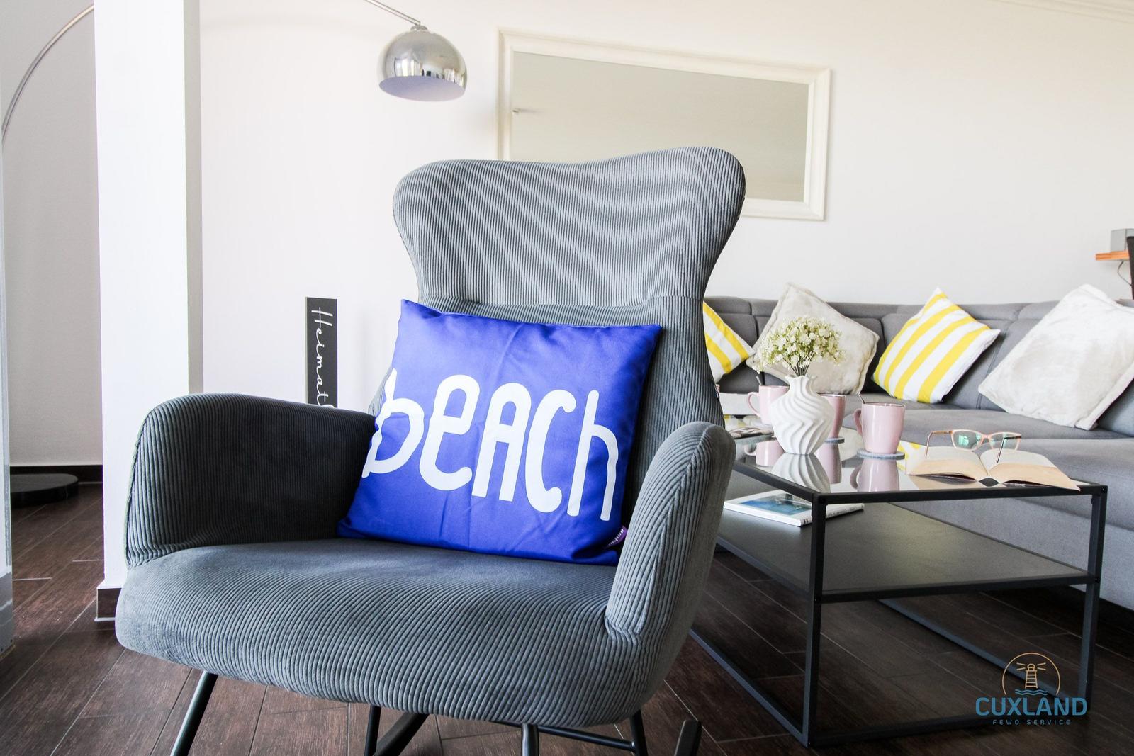 Graue Sitzgelegenheit mit blauem Kissen und Sofa im Hintergrund.