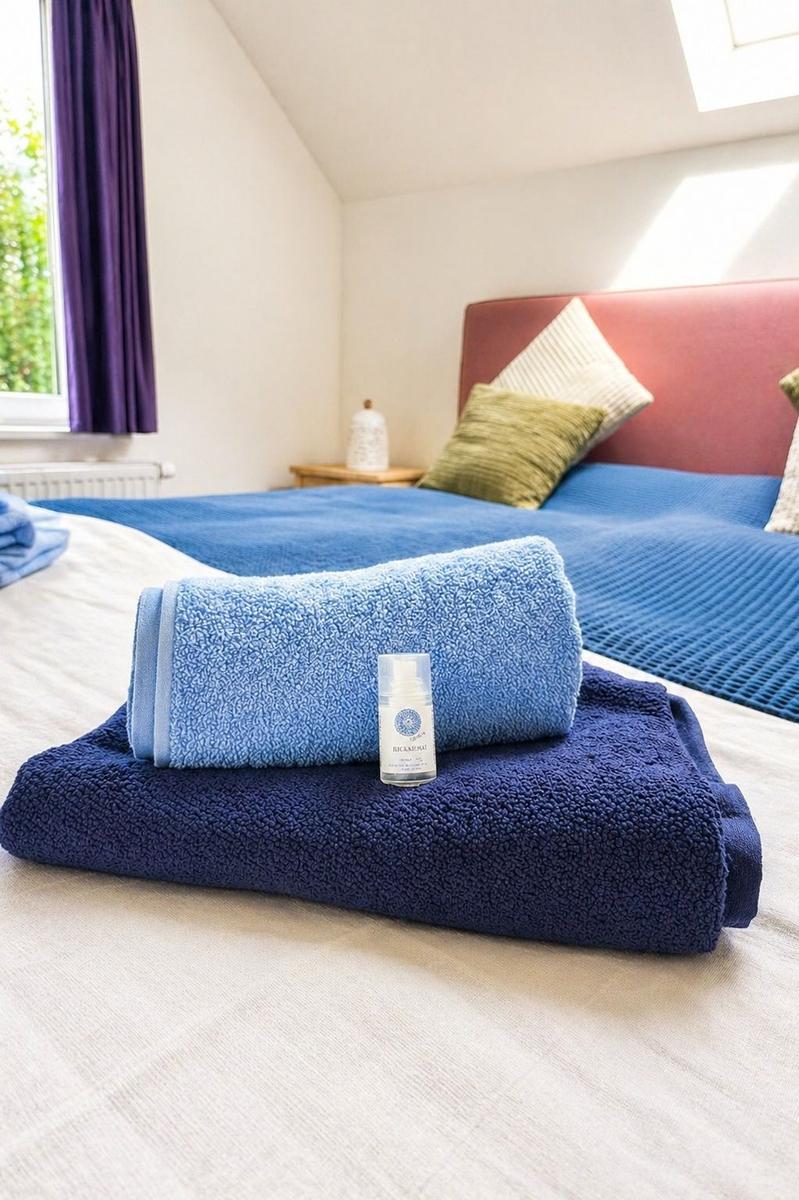 Ein Bett mit blauen Handtüchern und einem Fläschchen darauf. Im Hintergrund ein Fenster und ein Kopfteil.