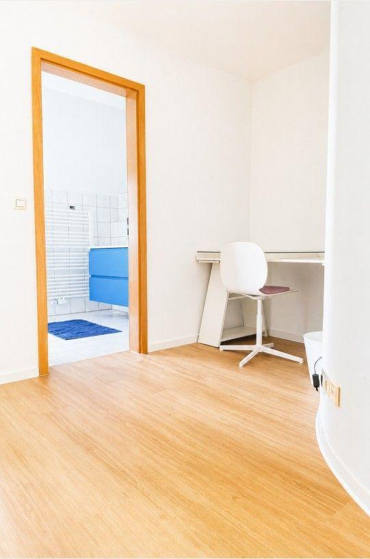 Ein Raum mit Holzboden, Schreibtisch und Stuhl. Durch Tür ist Bad mit blauem Waschbecken sichtbar.