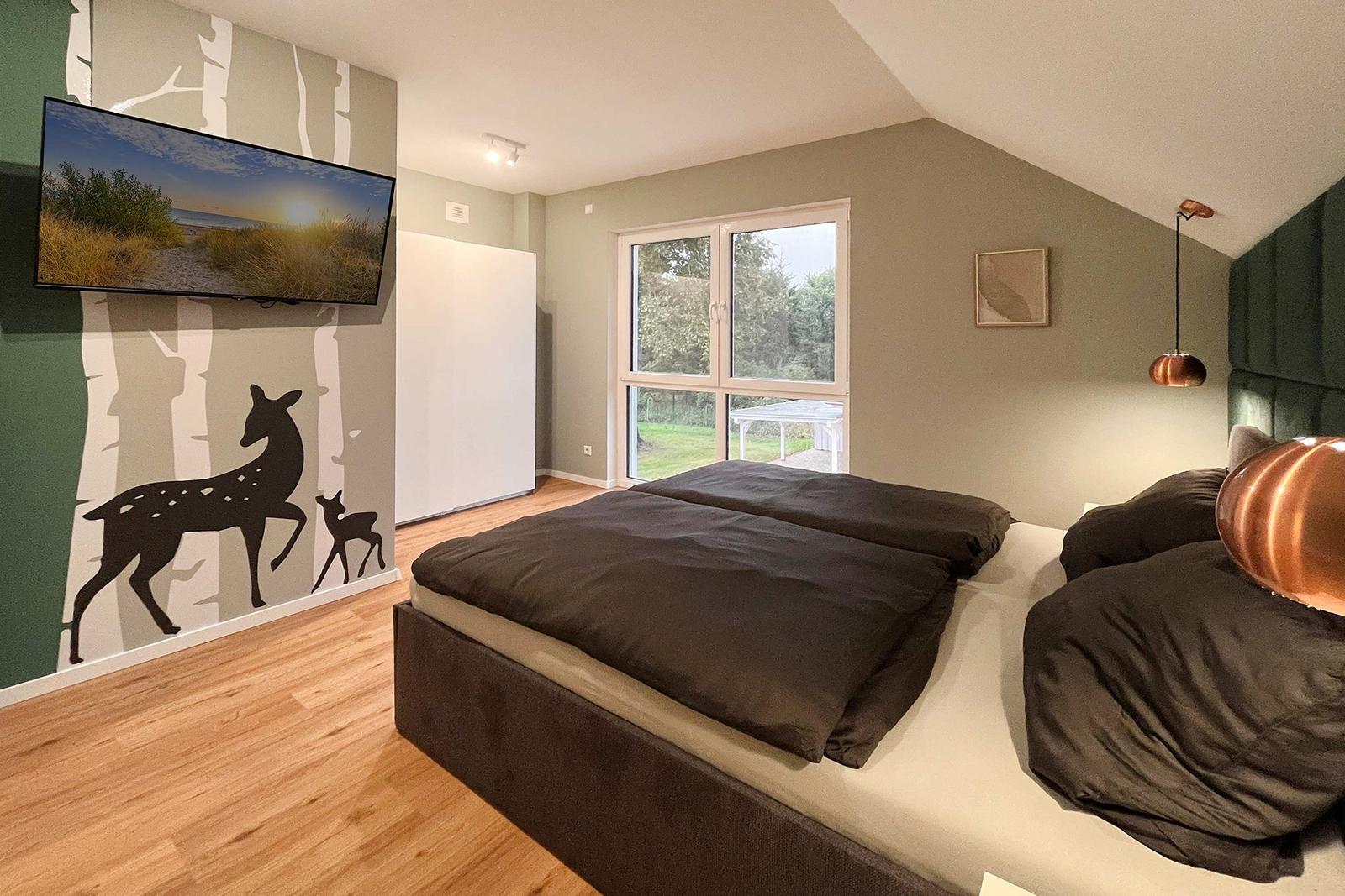 Schlafzimmer mit Bett, Fenster und Wandbild mit Hirsch. Holzboden und Deckenleuchte.