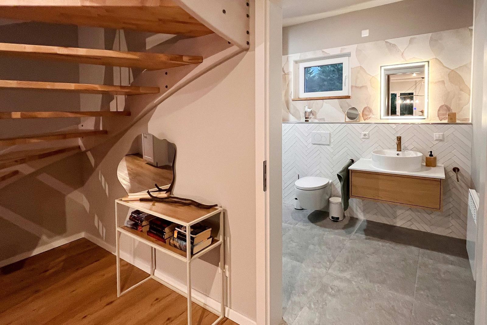Badezimmer mit Waschbecken, Toilette und Fenster. Holztreppe und kleiner Tisch mit Büchern.