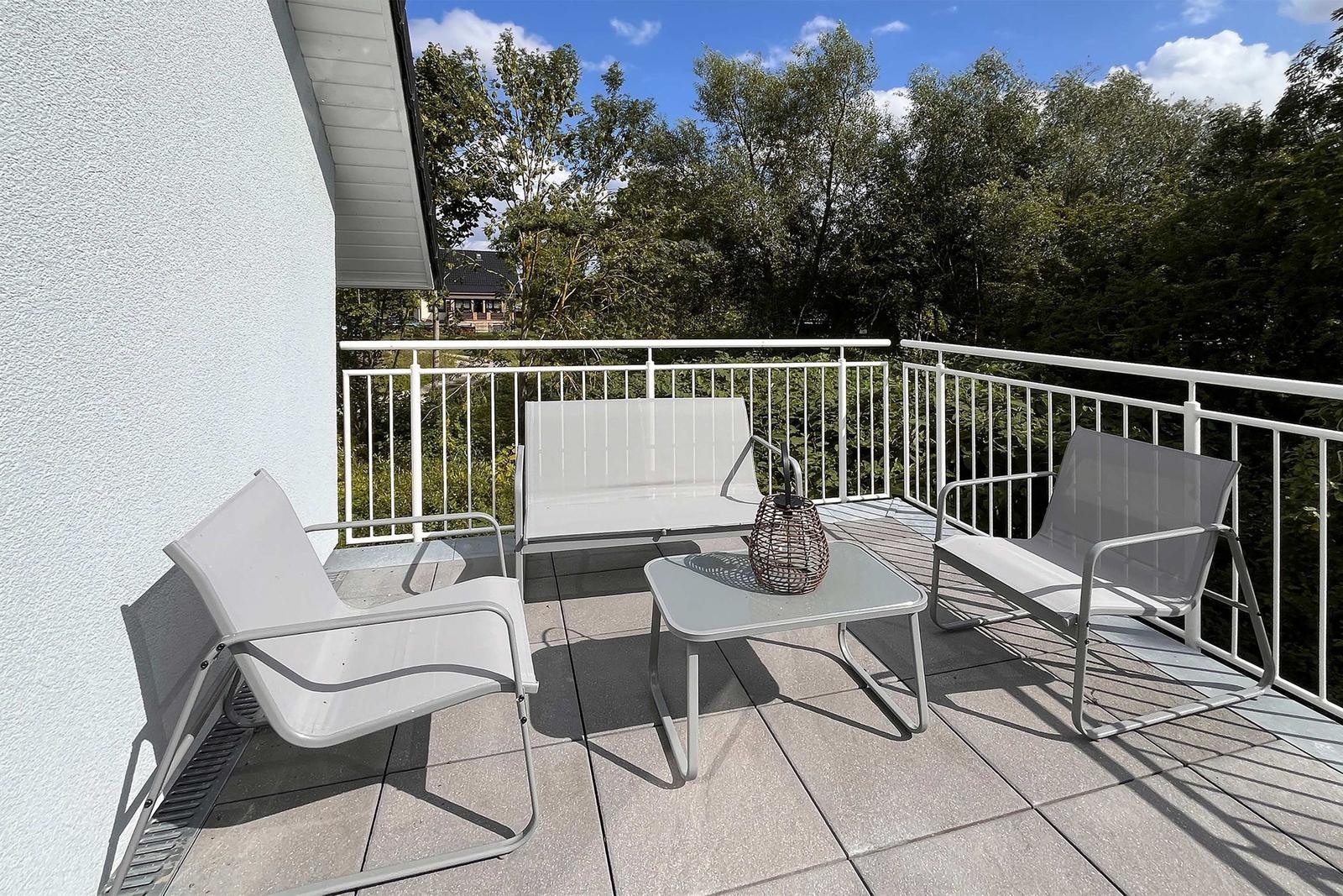 Terrasse mit weißen Sitzmöbeln und Tisch. Hintergrund: grüne Bäume und blauer Himmel.