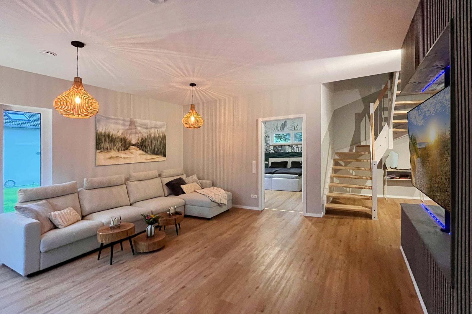 Wohnzimmer mit Sofa, Tischen und Treppenaufgang. Fernseher und Schlafzimmer sichtbar.