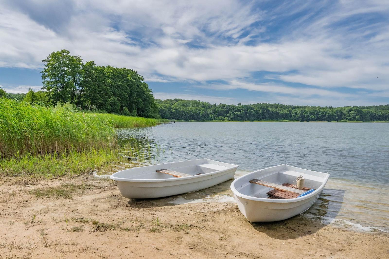 Zwei weiße Boote stehen am Strand eines Sees mit grüner Vegetation und Wald im Hintergrund.