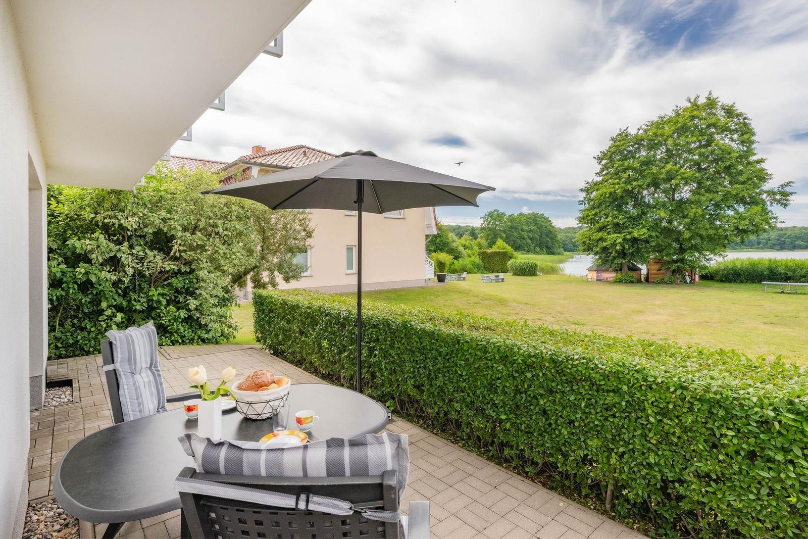Terrasse mit Tisch, Stühlen und Sonnenschirm. Blick auf Rasen, Hecke und Wasser.