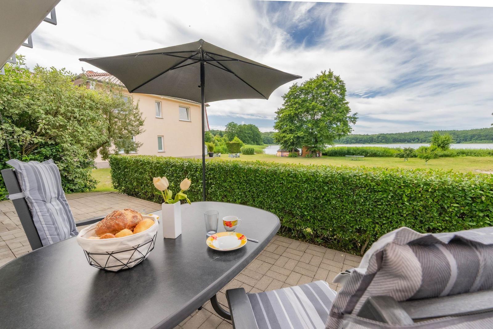 Terrasse mit Tisch, Stühlen und Sonnenschirm. Blick auf Grünfläche und Wasser.