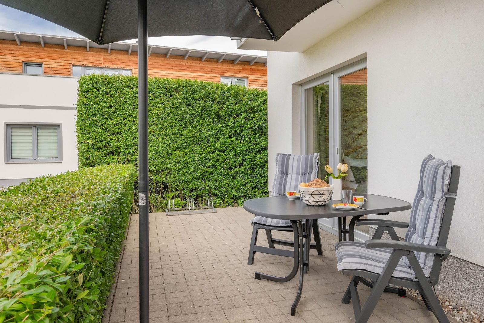 Terrasse mit Tisch, zwei Stühlen und Sonnenschutz. Hintergrund: grüner Heckenzaun und Hauswand.