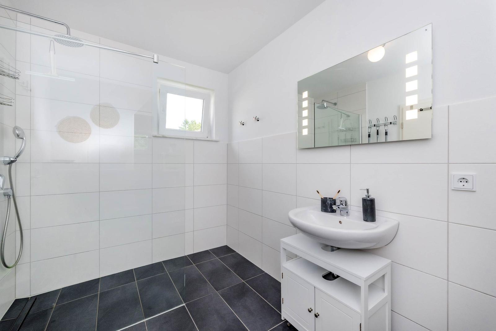 Badezimmer mit Dusche, Waschbecken und Spiegel. Weiße Wände, dunkle Bodenfliesen.