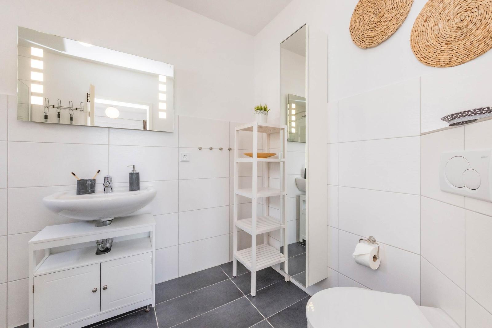 Badezimmer mit Waschbecken, Spiegel, Regal und Toilette. Weiße Fliesen und graue Bodenfliesen.