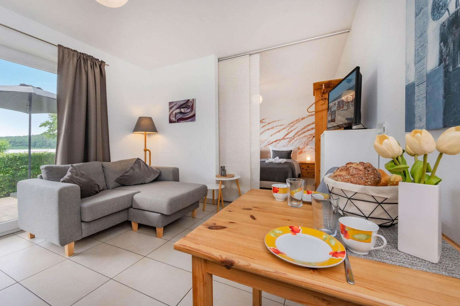 Wohnbereich mit Sofa, Tisch und Blick auf Balkon. Schlafzimmer im Hintergrund.