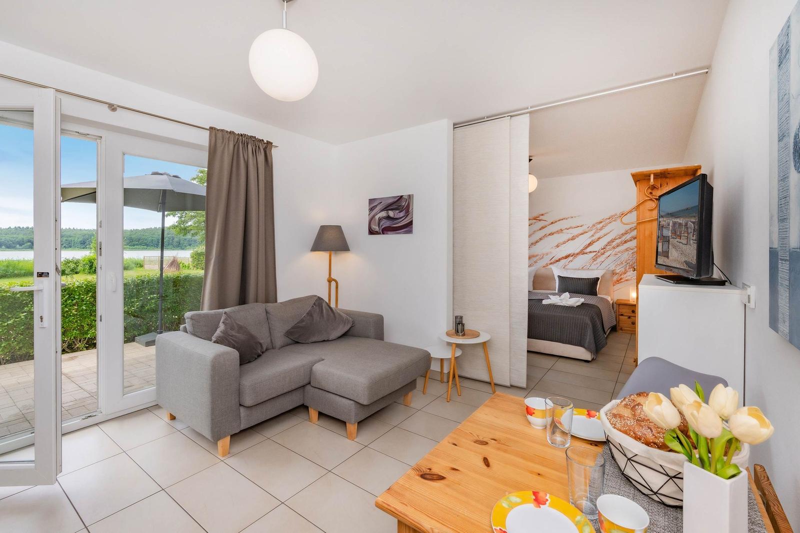 Wohnbereich mit Sofa, Couchtisch und Blick auf Terrasse. Schlafzimmer mit Bett und TV sichtbar.