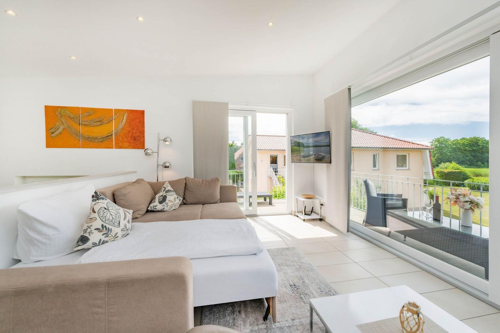 Wohnzimmer mit Sofa, Fernseher und Blick auf Balkon mit Gartenansicht.