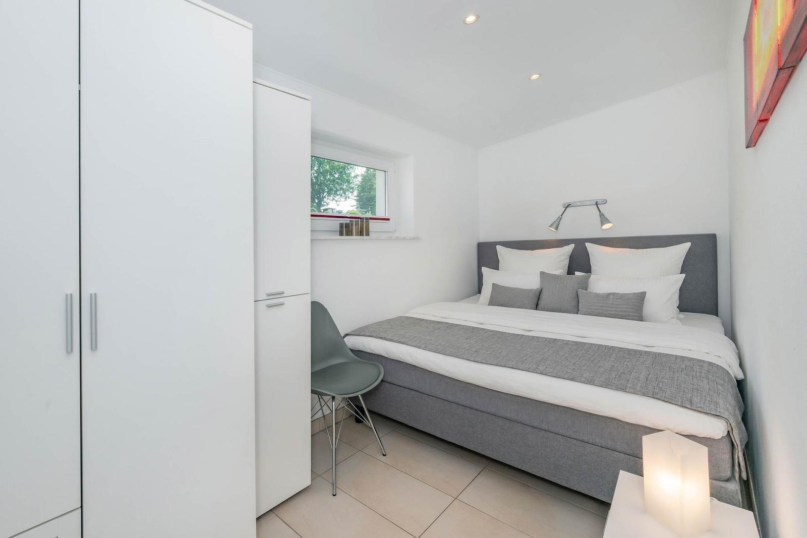 Schlafzimmer mit grauem Bett, weißen Schränken und kleinem Fenster.
