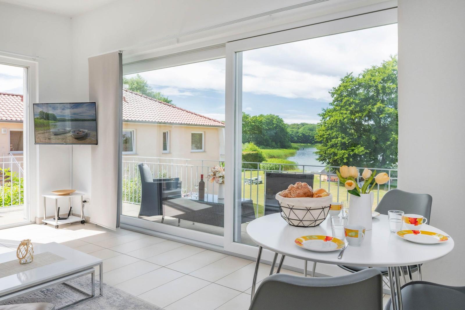 Essbereich mit Blick auf Balkon und See. Fernseher, Tisch und Stühle sind sichtbar.