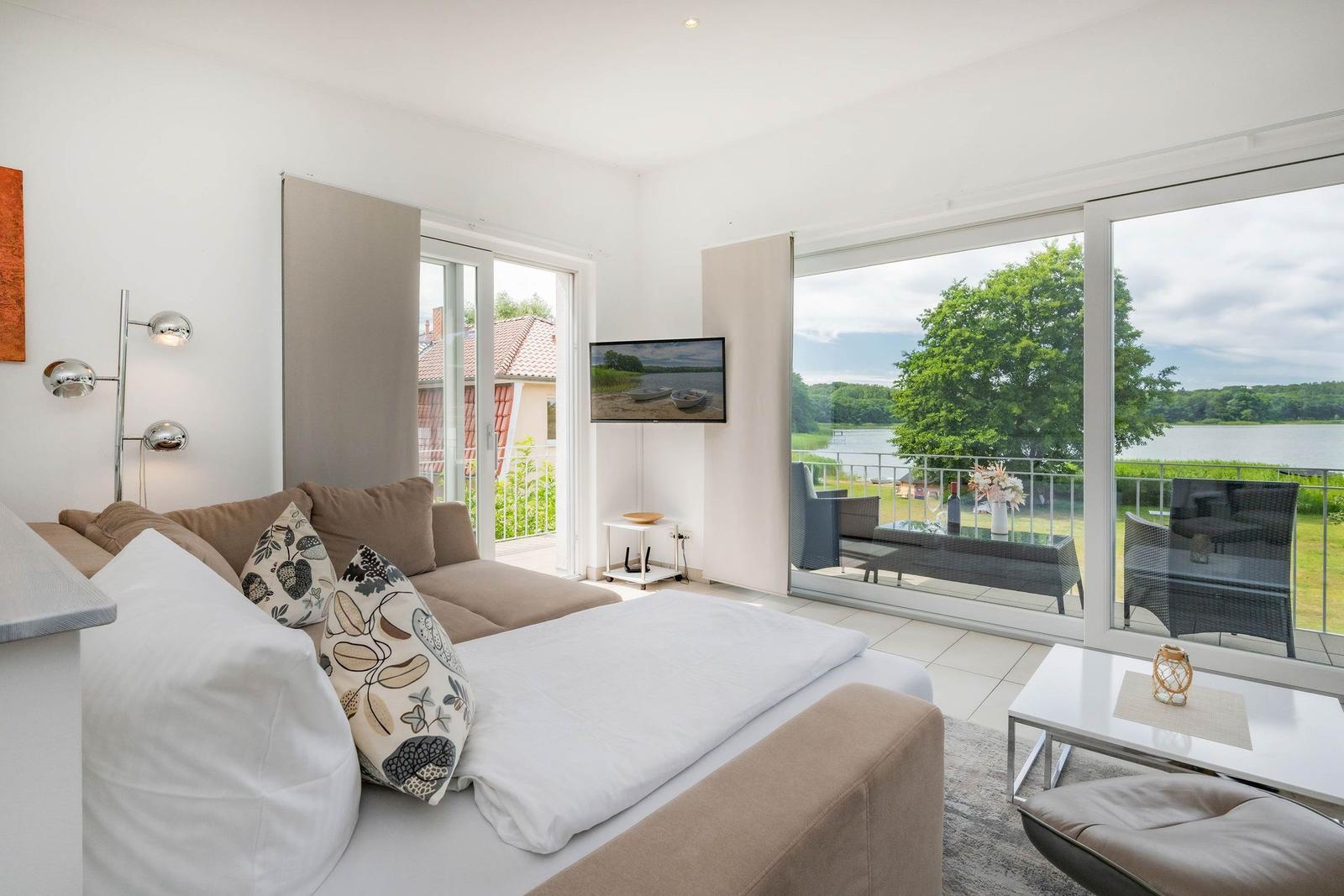 Wohnraum mit Blick auf den See und Balkon mit Sitzgelegenheiten.
