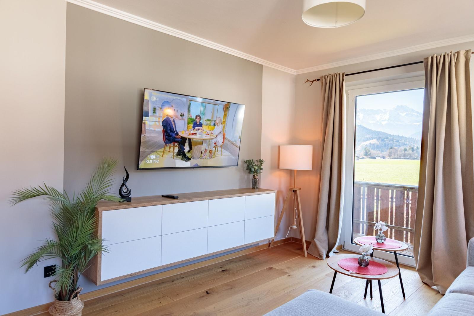 Wohnzimmer mit TV, Balkon und Bergblick. Holzboden, Couch und dekorative Pflanzen.