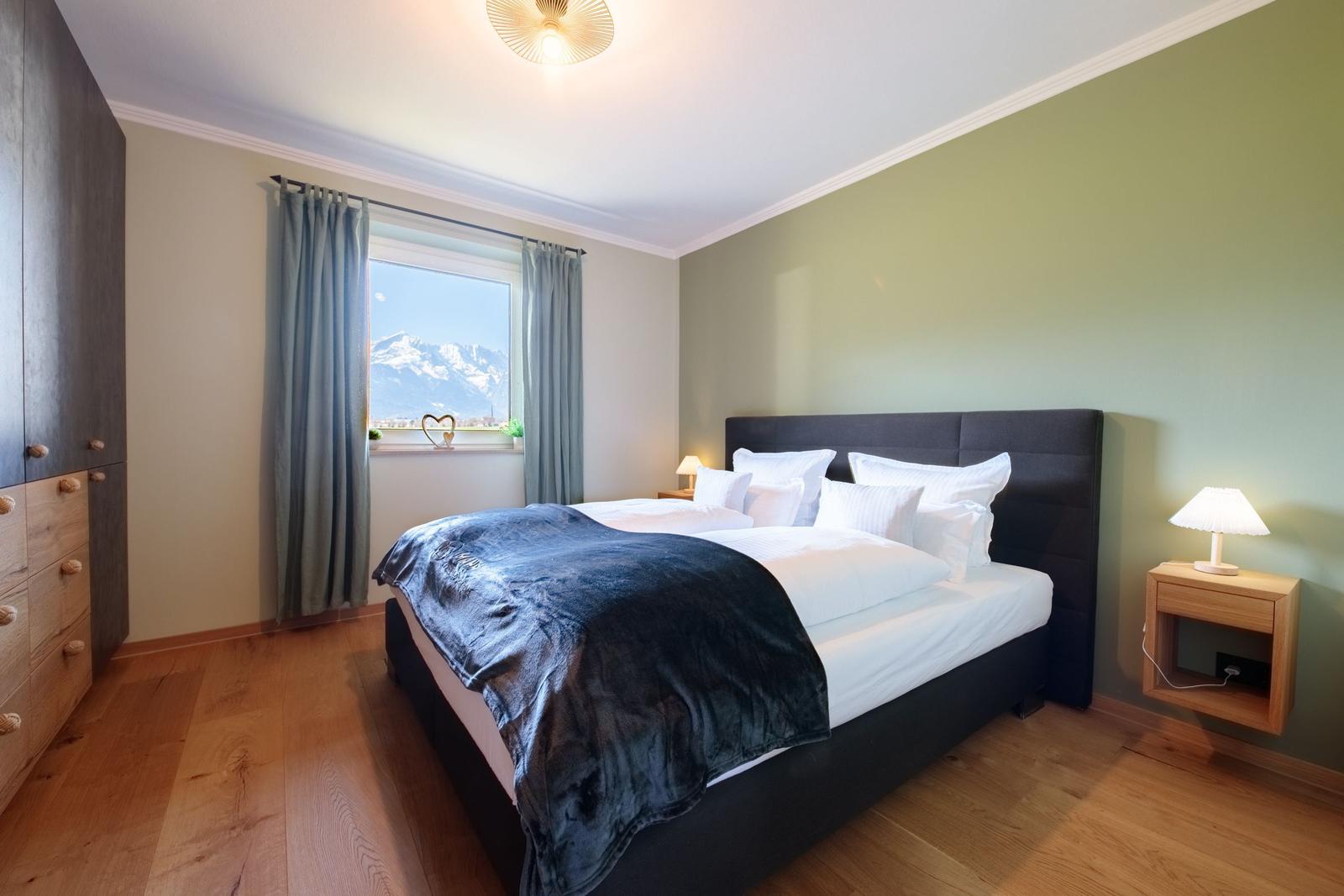 Schlafzimmer mit Bett, Nachttisch und Fensterblick auf Berge.