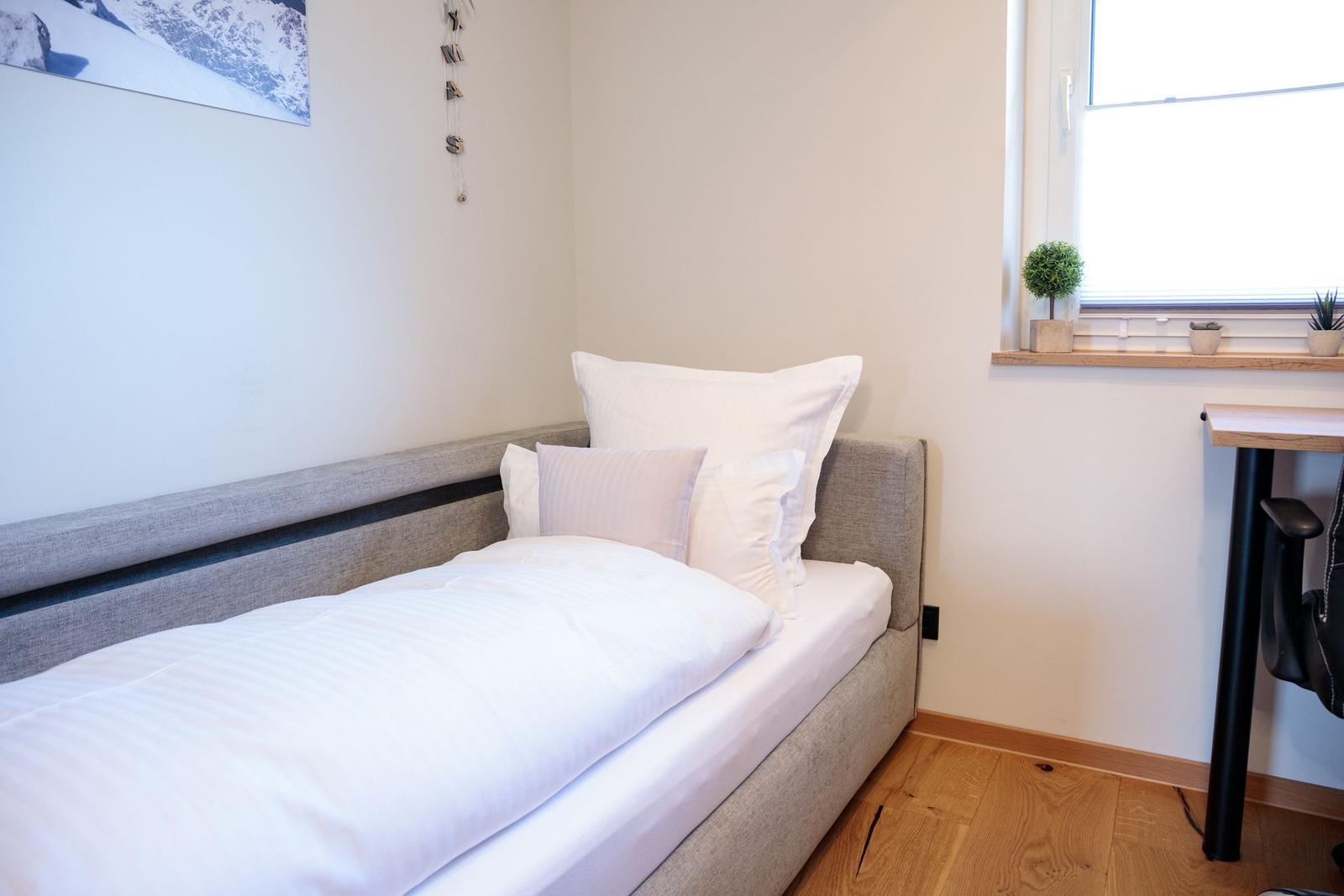 Ein Schlafzimmer mit Bett, Fenster und Schreibtisch. Wandbild und Pflanzen sind sichtbar.