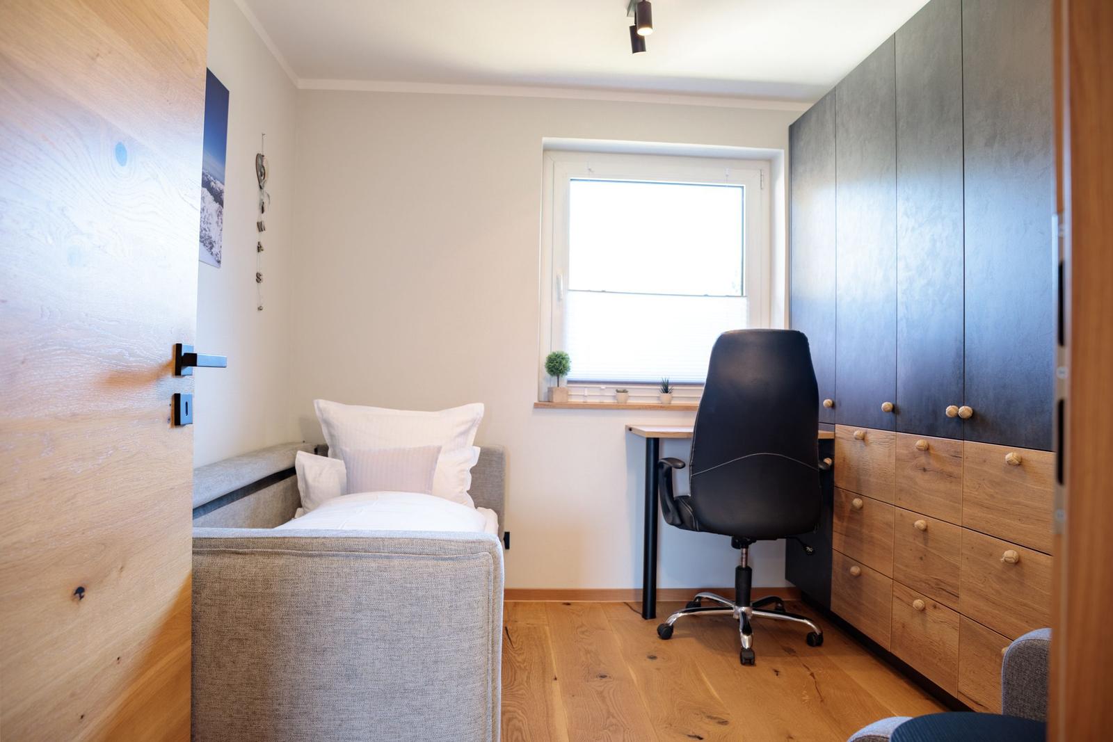 Schlafzimmer mit Bett, Schreibtisch und Schrank. Holzboden und Fenster mit Pflanzen.