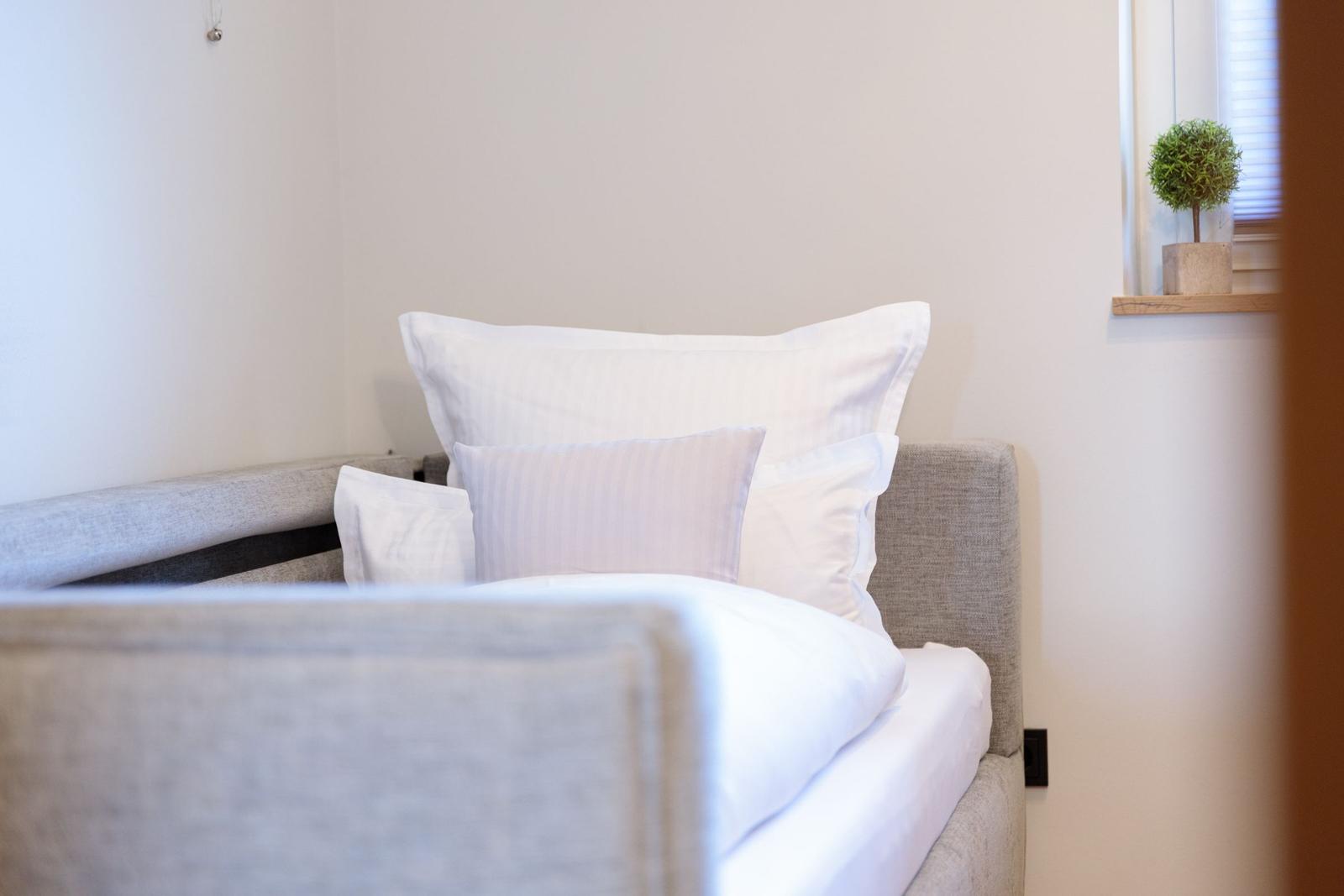 Ein Bett mit weißen Kissen und Bettwäsche. An der Wand hängt ein kleiner Topf mit Pflanze.