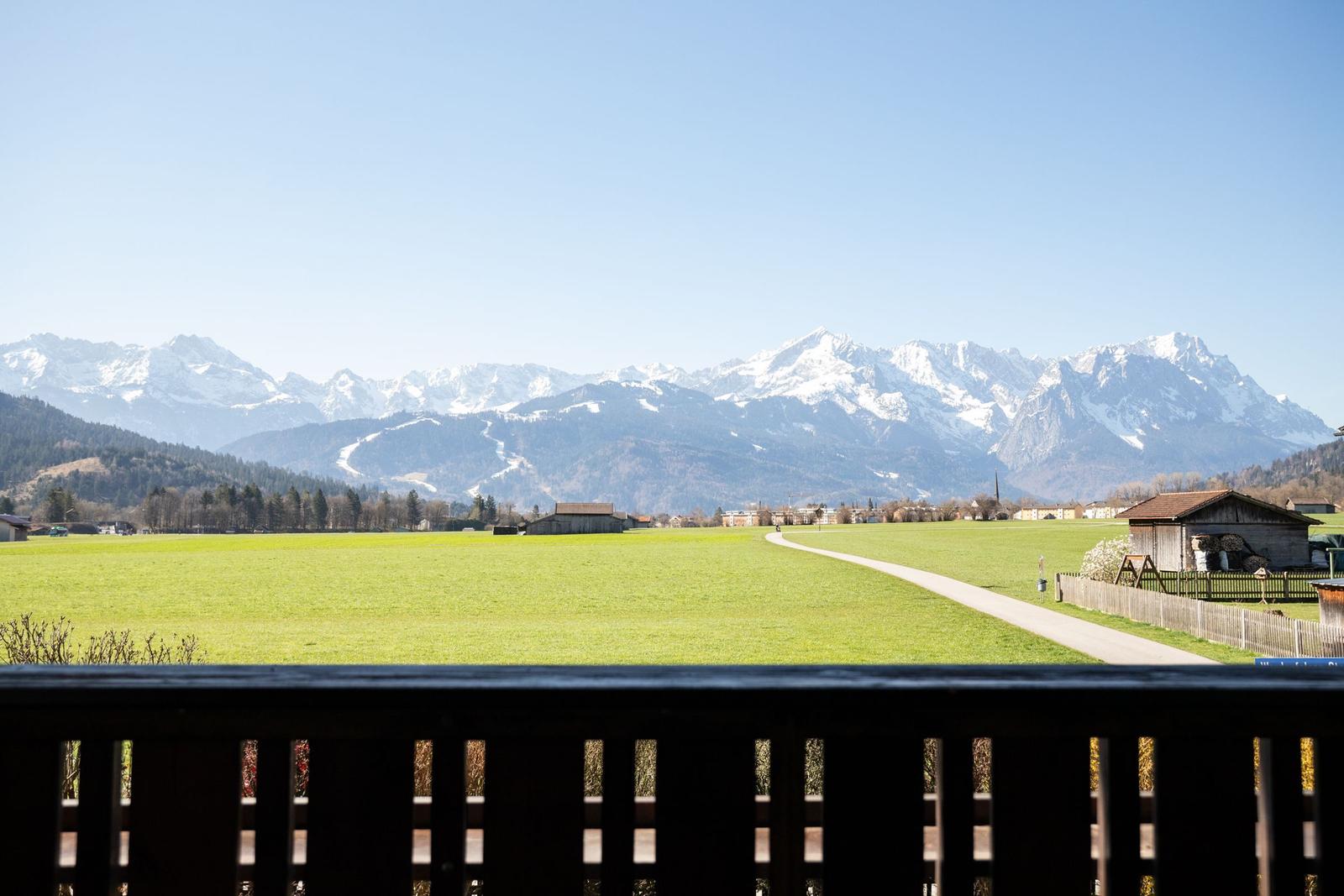 Blick von Balkon auf grünes Feld, Weg und schneebedeckte Berge.