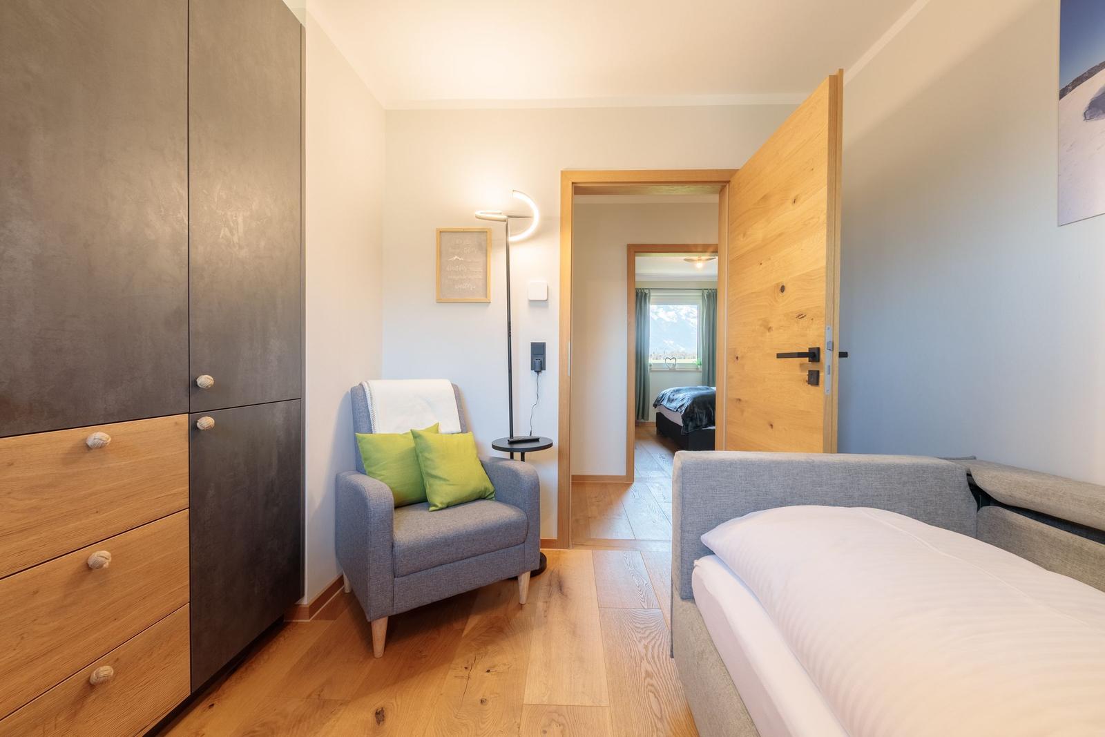 Schlafzimmer mit Bett, Sessel und Kleiderschrank. Tür führt in Schlafbereich mit Blick.