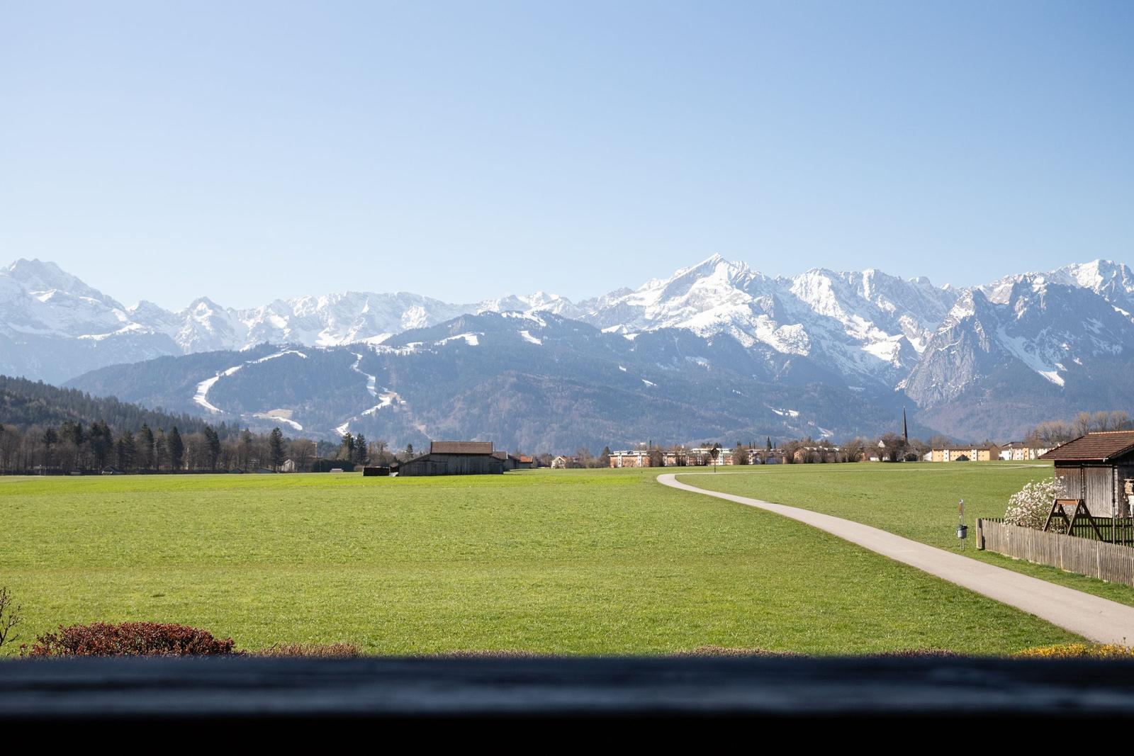 Weitläufiges Grünfeld mit Pfad, dahinter Berglandschaft mit Schneekappen und Siedlung.