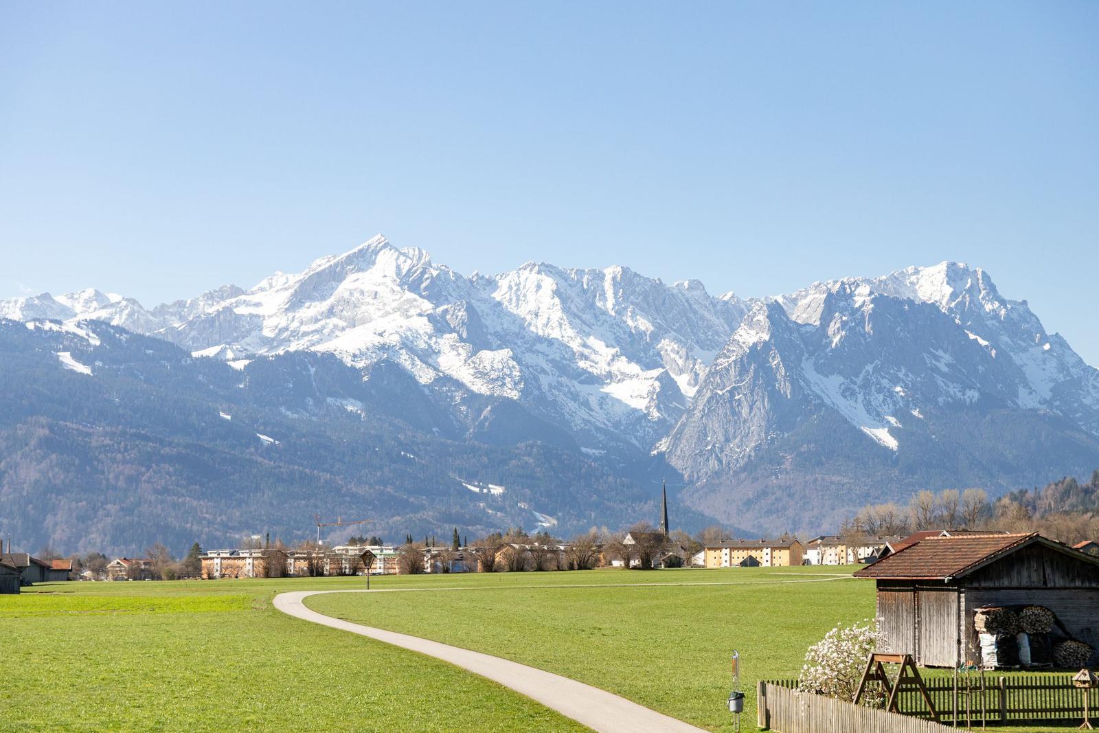 Weitläufiges Feld mit Pfad, Dorf und schneebedeckten Bergen im Hintergrund.