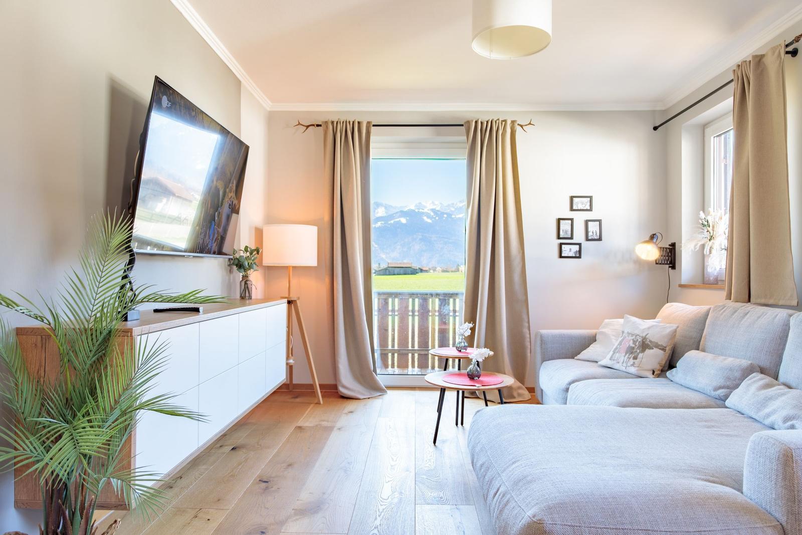 Wohnzimmer mit Sofa, Fernseher und Balkonblick auf Berge.