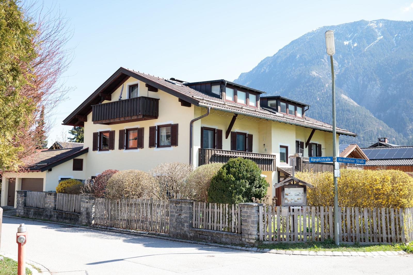 Ein zweistöckiges Haus mit Balkon und Dachfenstern, umgeben von einem Zaun und Bergen im Hintergrund.