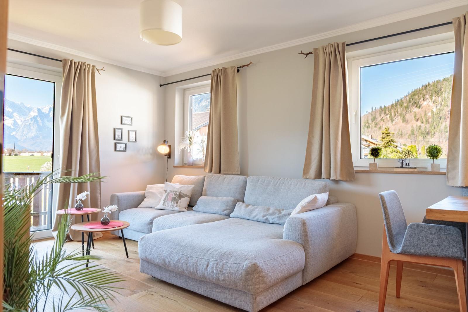 Wohnzimmer mit Sofa, Tisch und Blick auf Berge. Holzboden und Fenster mit Vorhängen.