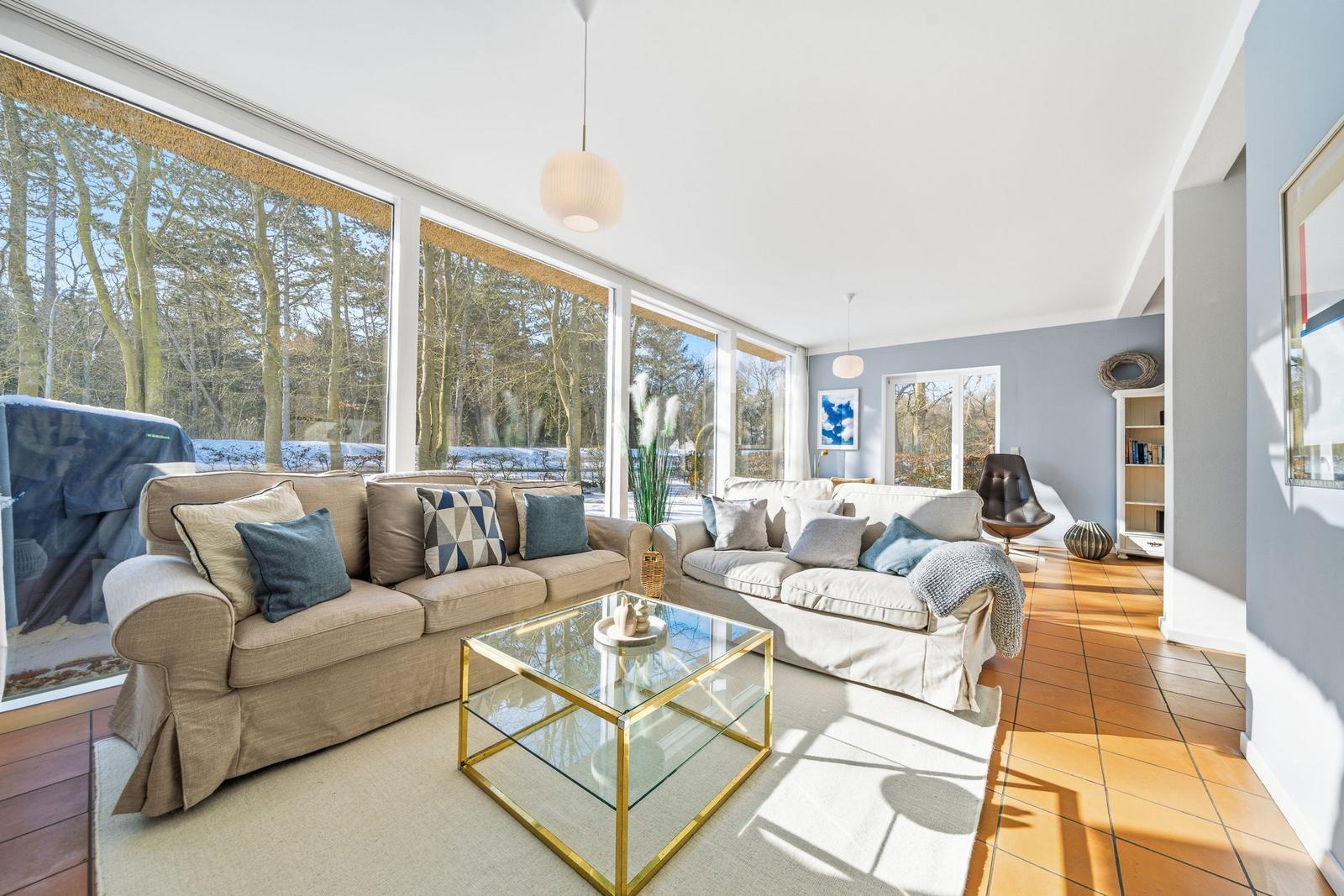 Wohnzimmer mit zwei Sofas, Glas-Tisch und großem Fensterfront zum Wald.