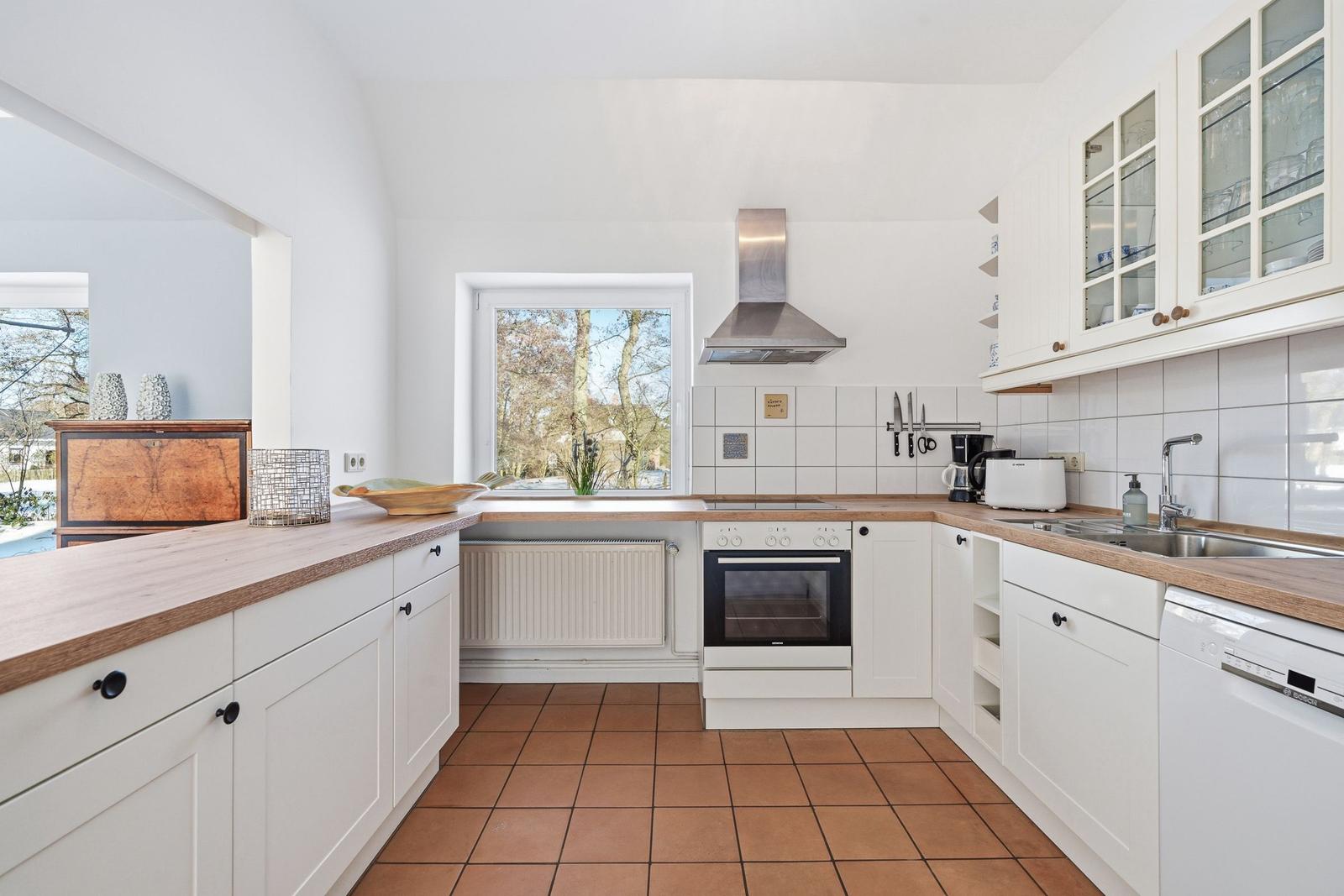 Küche mit weißen Schränken, Holzoberflächen und Terrakottboden. Fenster mit Blick auf Außenbereich.