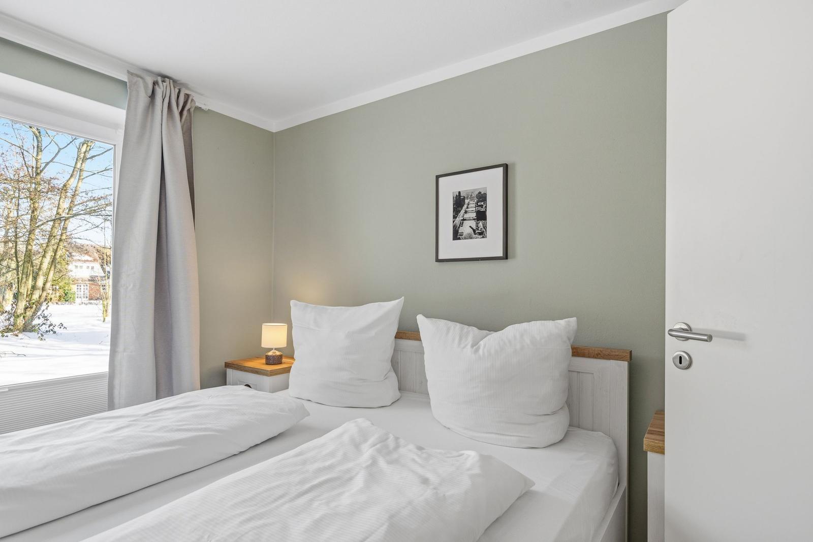 Schlafzimmer mit Doppelbett, Fenster mit Blick auf Schnee und Bäume, Nachttisch mit Lampe und Wandbild.