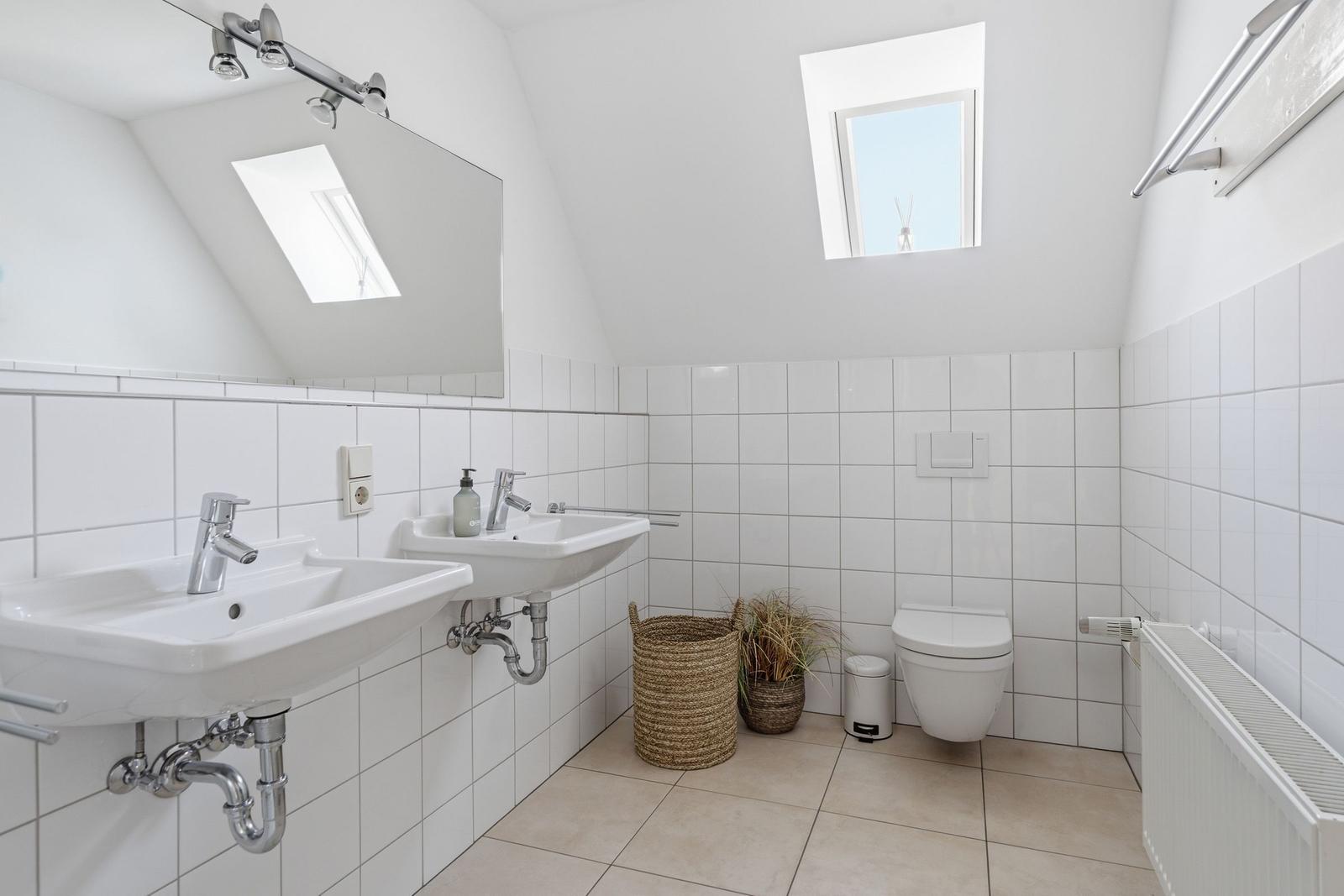 Badezimmer mit zwei Waschbecken, Toilette und Dachfenster. Weiße Fliesen und Holzdielen.