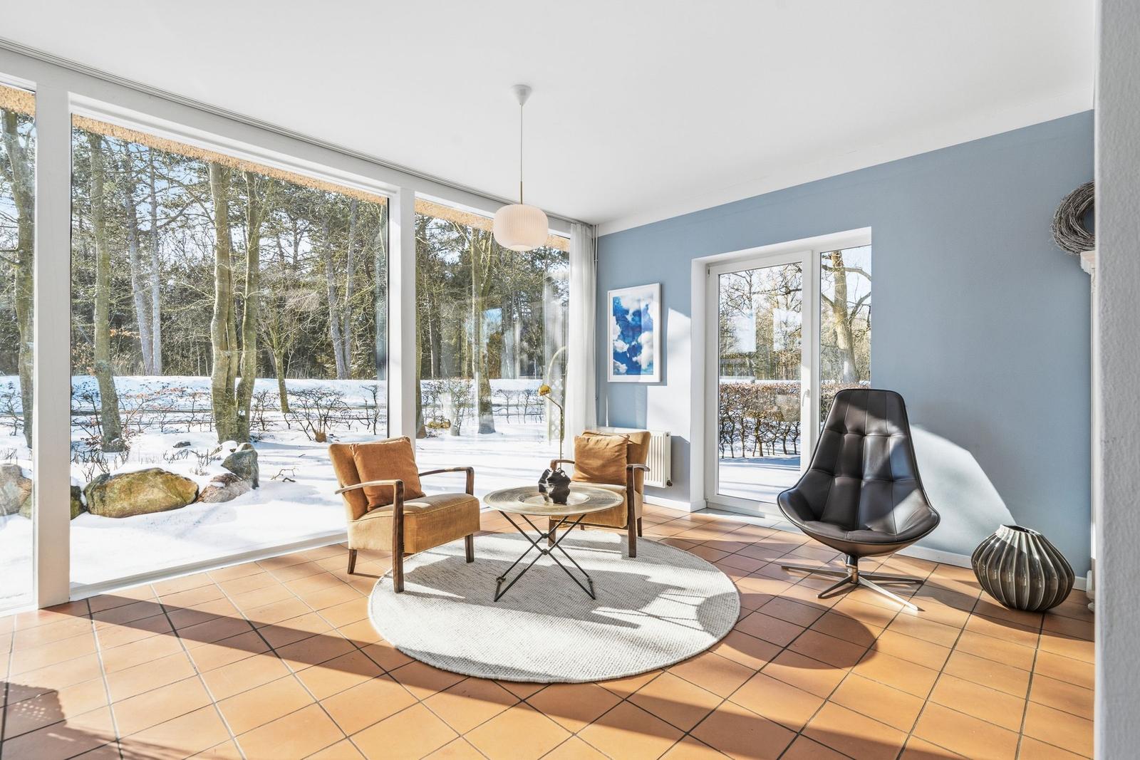 Ein Wohnzimmer mit großen Fenstern, zwei Sesseln und einem runden Teppich auf Terrakottaboden.