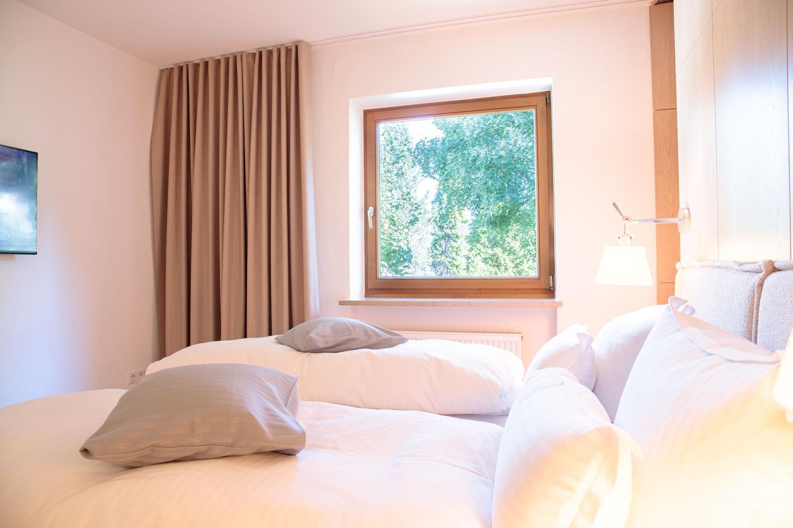Ein Schlafzimmer mit Bett, Fenster und Vorhang. Draußen sind Bäume sichtbar.