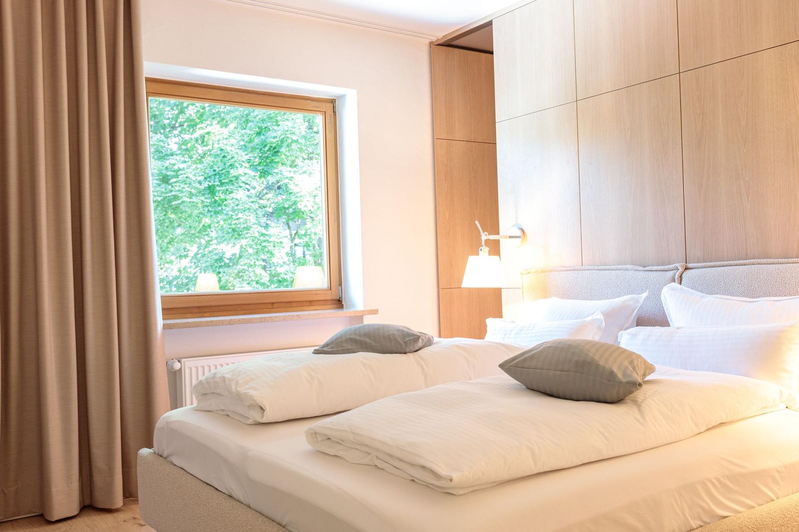 Doppelbett mit weißen Bettwäsche und grauen Kissen. Holzfenster mit Blick auf Bäume.