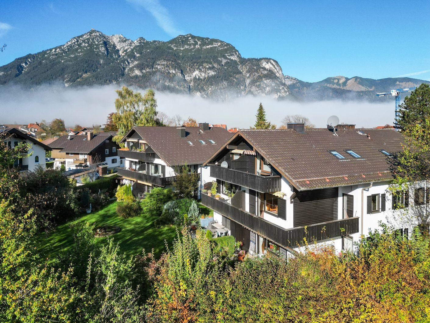 Haus mit Balkon und Garten. Berge im Hintergrund. Nebel liegt zwischen den Bergen.