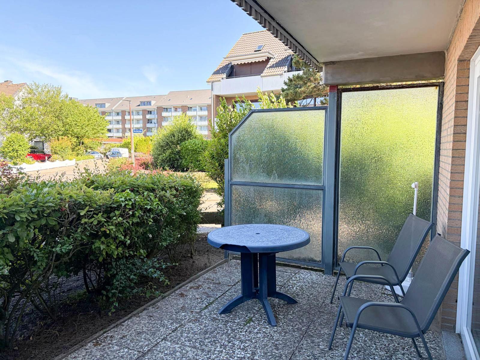 Terrasse mit Tisch und zwei Stühlen, umgeben von Grünflächen und einem Glaswandschutz.