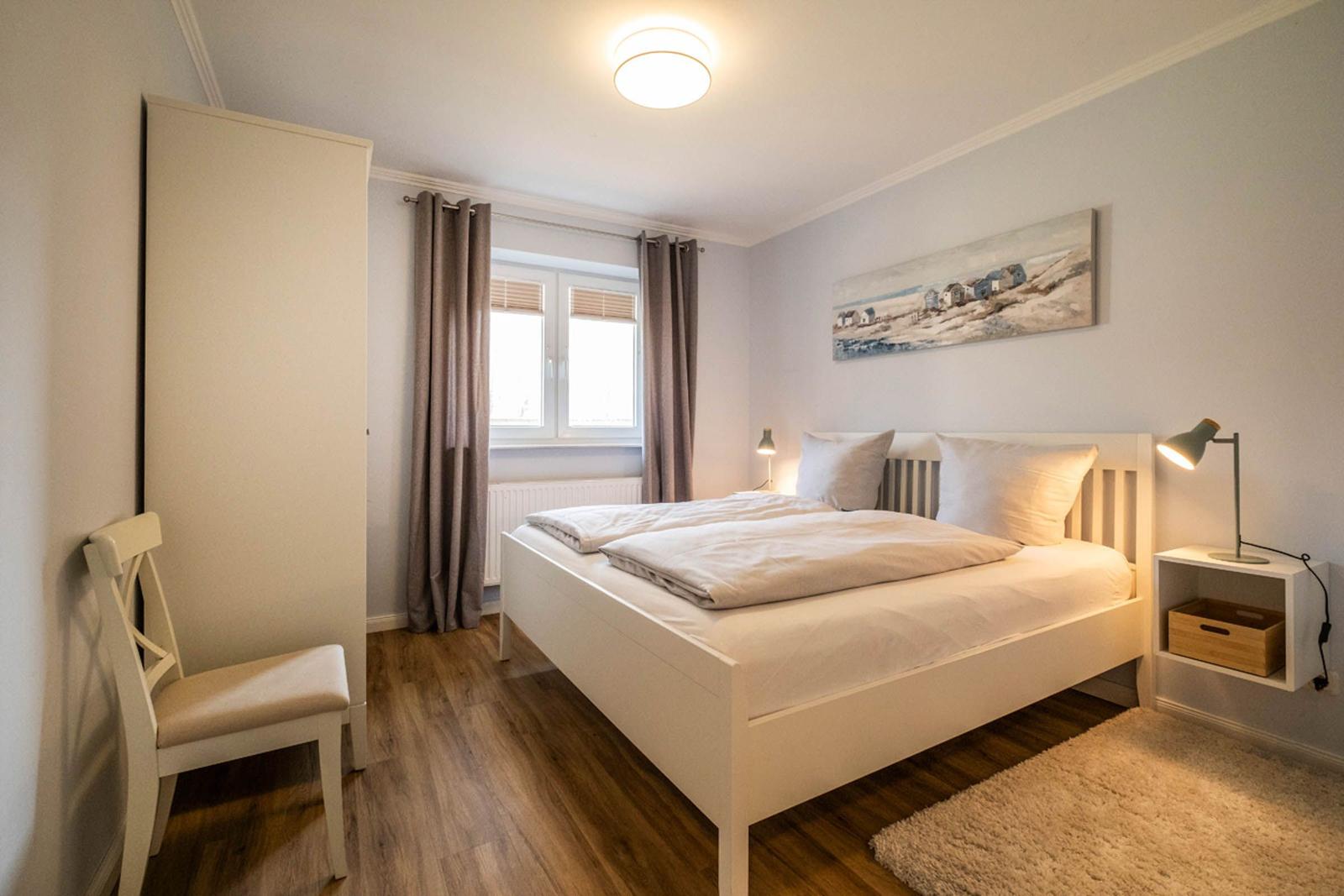 Ein Schlafzimmer mit weißem Bett, Holzboden und Fenster mit Vorhängen.